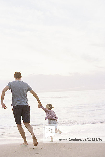 Man playing on a sandy beach by the ocean  holding his young daughter's hand.