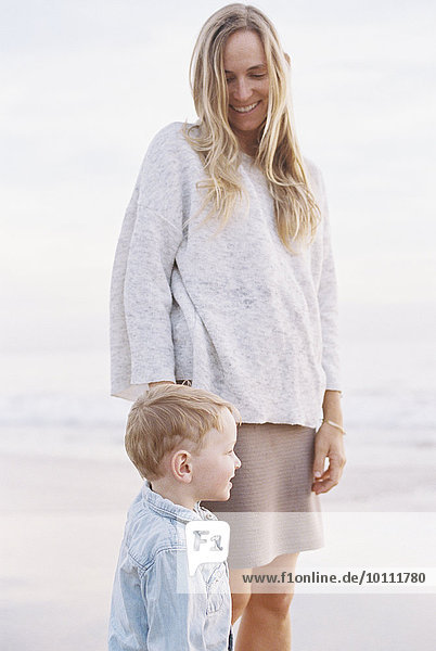 Smiling woman standing on a sandy beach by the ocean with her young son.