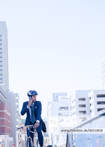 Geschäftsmann im Anzug und Helm im Gespräch auf dem Handy auf dem Fahrrad in der Stadt