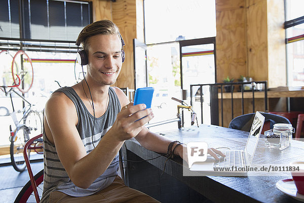 Lächelnder junger Mann mit Kopfhörer SMS mit Handy am Laptop im Cafe