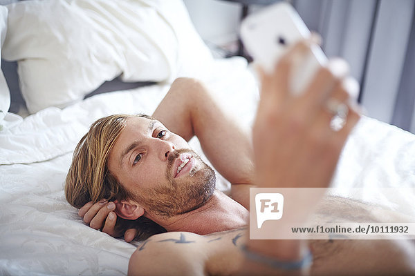 Mann mit nackter Brust auf dem Bett liegend SMS mit Handy