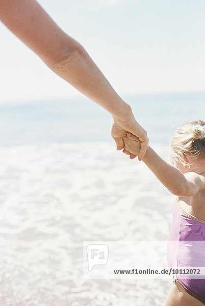 Woman and her daughter holding hands  standing on a sandy beach by he ocean.