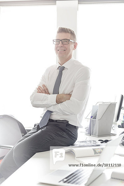 Portrait selbstbewusster Geschäftsmann mit Brille auf dem Schreibtisch sitzend mit gekreuzten Armen