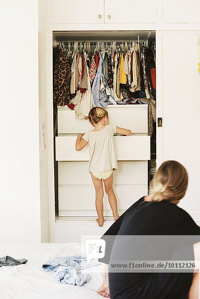 Young girl looking for clothes in a chest of drawers  her mother sitting on a bed  watching.