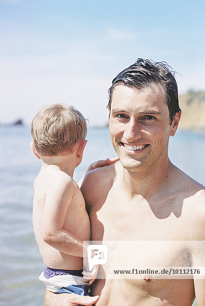 A man on the beach  smiling  carrying his young son