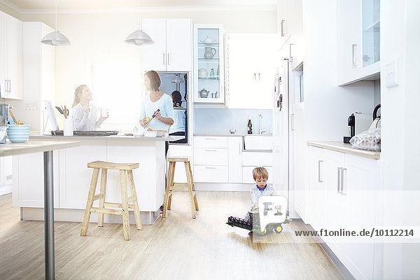 Women cooking in kitchen while boy plays with toy tractor on floor