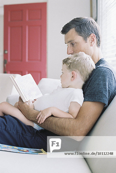Man sitting on a sofa with his son sitting on his lap  holding a book  reading a story.