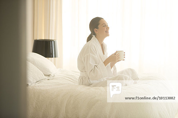 Smiling woman in bathrobe drinking coffee cross-legged on bed