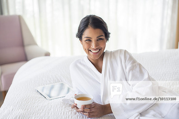 Portrait smiling woman in bathrobe drinking tea on bed