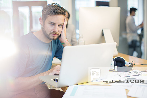 Serious creative businessman using laptop with head in hands in office