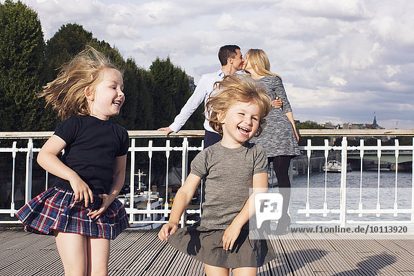 Kleine Mädchen spielen im Freien,  während ihre Eltern sich im Hintergrund küssen.