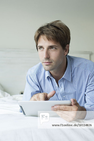Mann im Bett liegend mit digitalem Tablett  wegblickend mit kontemplativem Blick