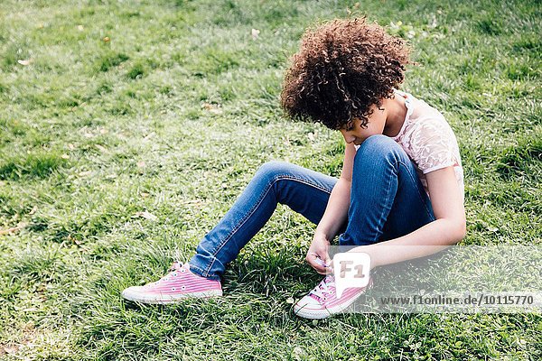 Mädchen in rosa Schuhen auf Gras sitzend  Schnürsenkel bindend  nach unten schauend
