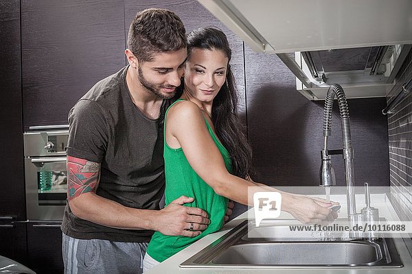 Junge Frau beim Abwaschen  während sie von ihrem Freund umarmt wird.