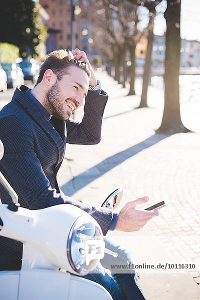 Stilvoller junger Mann sitzt auf Moped mit Smartphone