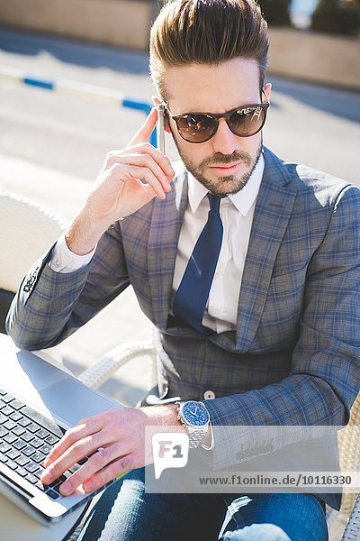 Stilvoller junger Geschäftsmann mit Smartphone und Laptop im Bürgersteigcafé