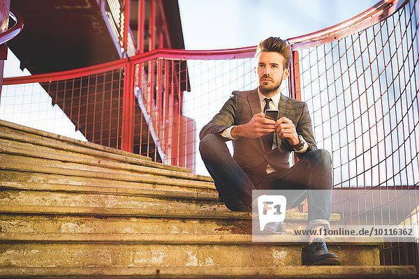 Stilvoller junger Mann sitzt auf Stegstreppe mit Smartphone