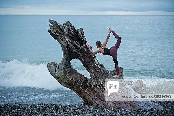 Reife Frau praktiziert Yogastellung auf großem Treibholzbaumstamm am Strand