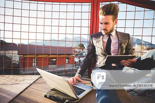 Stylish young man sitting on footbridge using laptop and digital tablet
