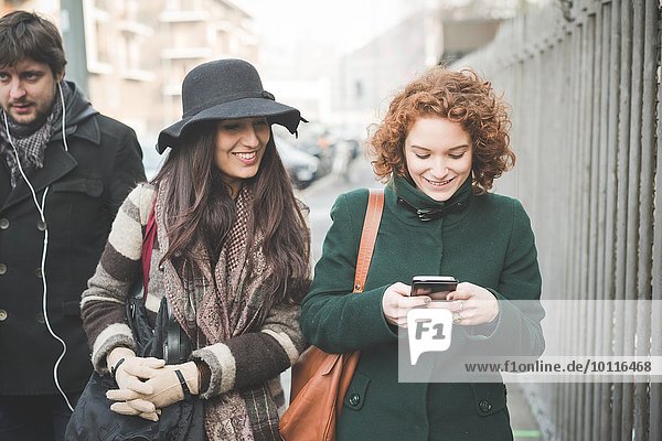 Two young women strolling along street reading smartphone texts
