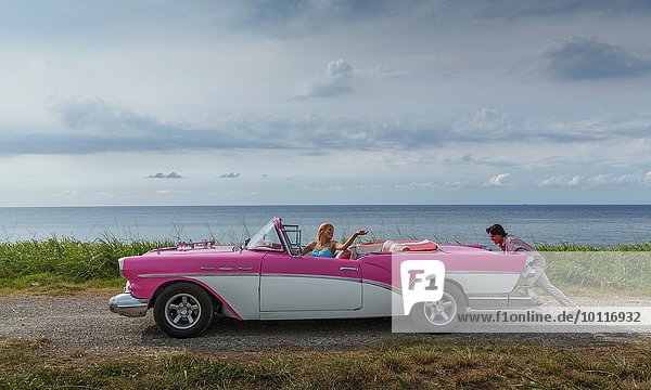 Junger Mann schiebt Vintage Cabrio an der Küste  während Freundin fährt  Havanna  Kuba