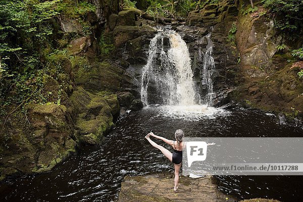 Rückansicht der reifen Frau  die vor dem Wasserfall Yoga praktiziert.