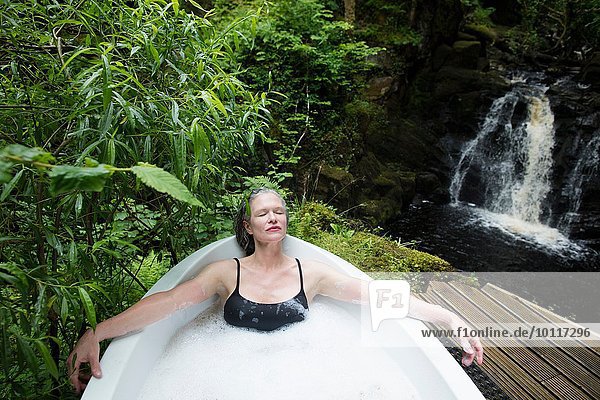 Reife Frau entspannt sich im Schaumbad vor dem Wasserfall beim Öko-Retreat