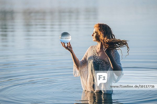 Junge Frau mit langen roten Haaren steht im See und schaut auf die Kristallkugel.