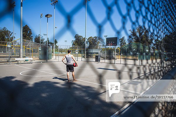 Young male basketball player behind court fence looking at basketball hoop