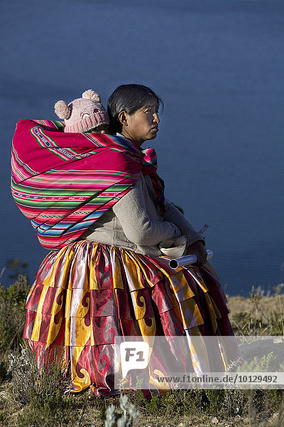 Indio woman with a young child on her back  traditional clothing  in front of lake Titicaca  Lake Titicaca  Sun Island  Isla del Sol  Bolivia  South America