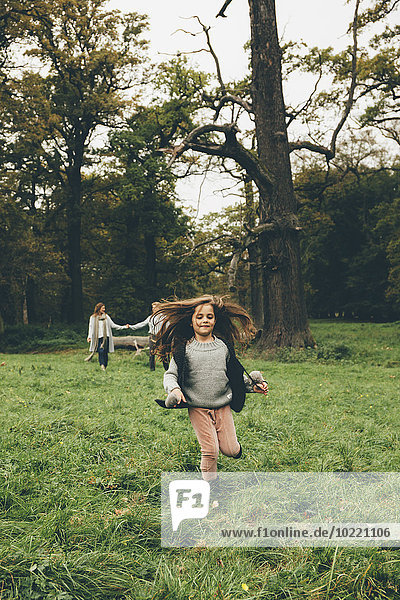 Kleines Mädchen läuft auf einer Wiese in einem Park  während ihre Eltern im Hintergrund stehen.