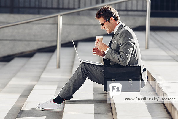 Geschäftsmann sitzt auf einer Treppe  hält einen Kaffee und arbeitet am Laptop.