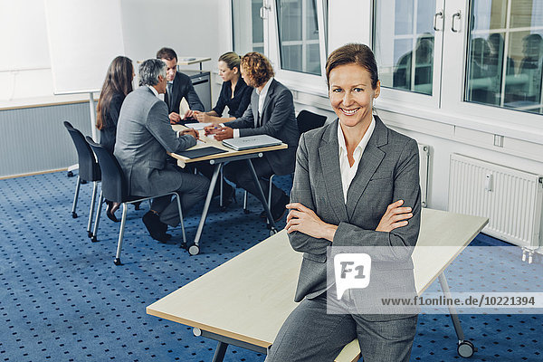 Reife Geschäftsfrau auf dem Schreibtisch sitzend mit gekreuzten Armen  Teamarbeit im Hintergrund