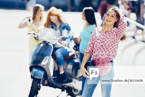 Porträt eines jungen Mädchens mit Motorradhelm  während ihre Freunde mit Roller im Hintergrund warten.