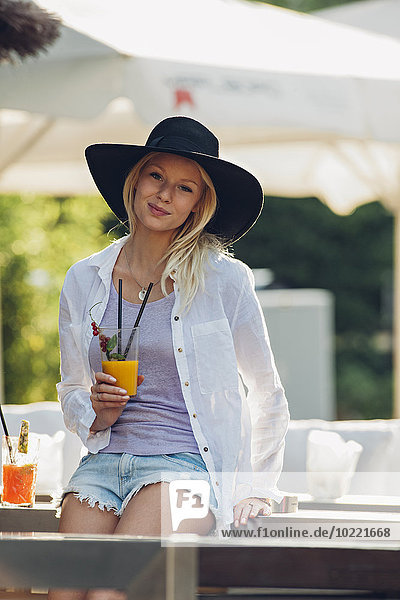 Portrait of young woman wearing hat sitting in an outdoor cafe with glass of orange juice