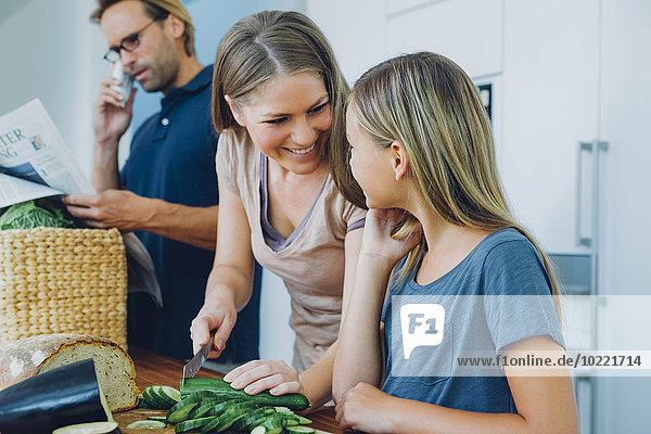 Mutter und Tochter in der Küche schneiden Gurke mit Vater im Hintergrund