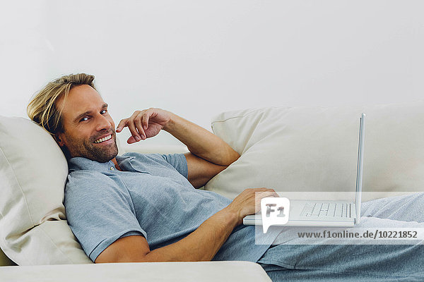 Smiling man lying on couch using laptop