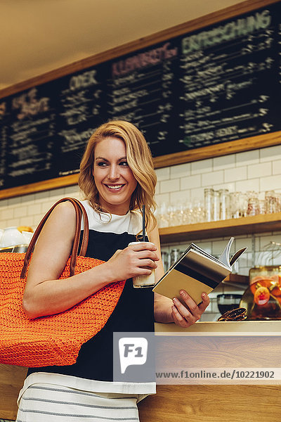 Portrait of smiling blond woman standing at counter in a coffee shop with booklet and Latte Macchiato