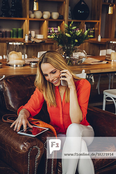 Blond woman with digital tablet sitting in a coffee shop telephoning with smartphone