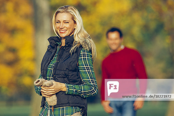 Porträt einer lächelnden  blonden Frau im herbstlichen Park mit einem Mann im Hintergrund.