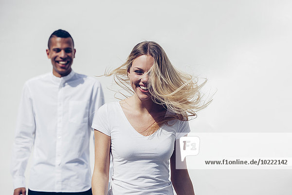Porträt einer lachenden  blonden Frau  die ihre Haare wirft  während ein junger Mann im Hintergrund steht.