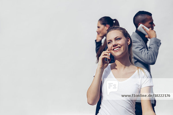 Porträt einer lächelnden jungen Frau  die mit dem Smartphone vor zwei anderen Menschen telefoniert.