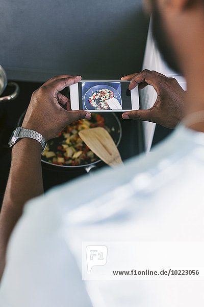 Junger Mann fotografiert sein zubereitetes Essen mit dem Smartphone in der Küche.