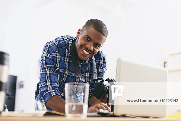 Porträt eines lächelnden jungen Mannes  der in seinem Fotostudio mit Laptop arbeitet.