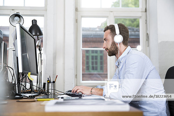Junger Mann im Büro mit Kopfhörer  der am Computer arbeitet