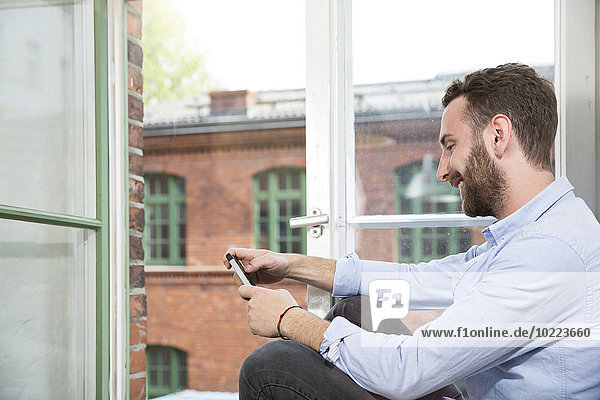Lächelnder junger Mann mit Handy am Fenster sitzend