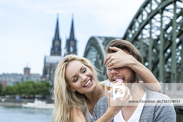 Germany  Cologne  young man tasting a bagel while his girlfriend covering his eyes