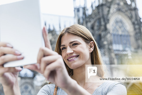 Deutschland  Köln  Porträt einer lächelnden jungen Frau  die vor dem Kölner Dom einen Selfie nimmt.
