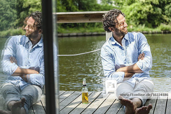 Lächelnder Mann sitzt auf der Plattform am Wasser mit Bierflasche