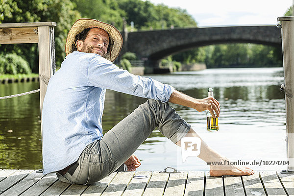 Lächelnder Mann sitzt auf der Plattform am Wasser mit Bierflasche